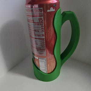 May include: A green 3D-printed can holder with a handle, designed to fit a standard soda can. The can is red with a nutrition facts label visible, including "Calories 15". The holder provides a comfortable grip.