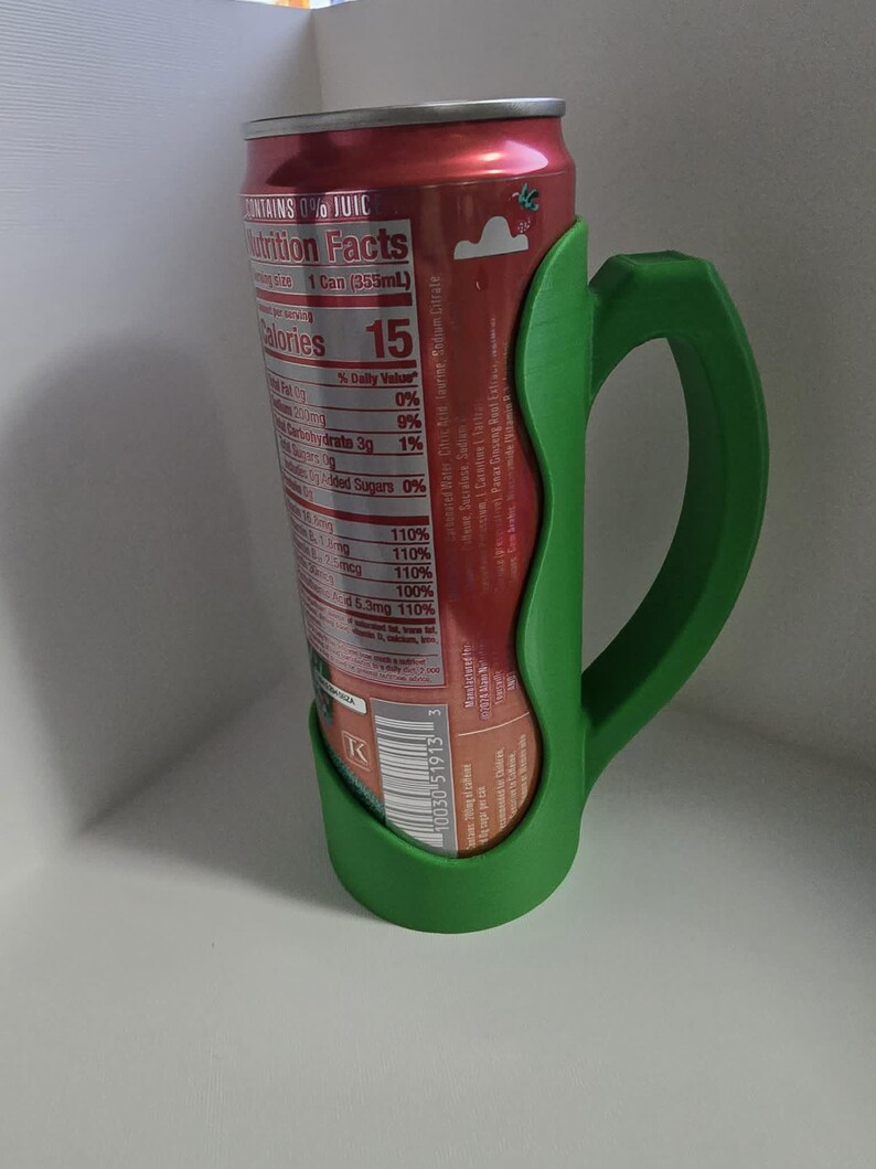 May include: A green 3D-printed can holder with a handle, designed to fit a standard soda can. The can is red with a nutrition facts label visible, including "Calories 15". The holder provides a comfortable grip.