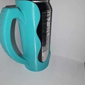 May include: A teal-colored can holder with a handle, designed to hold a standard-sized beverage can. The holder is made of a smooth, possibly 3D-printed material. A black can with a nutrition label is visible inside the holder.