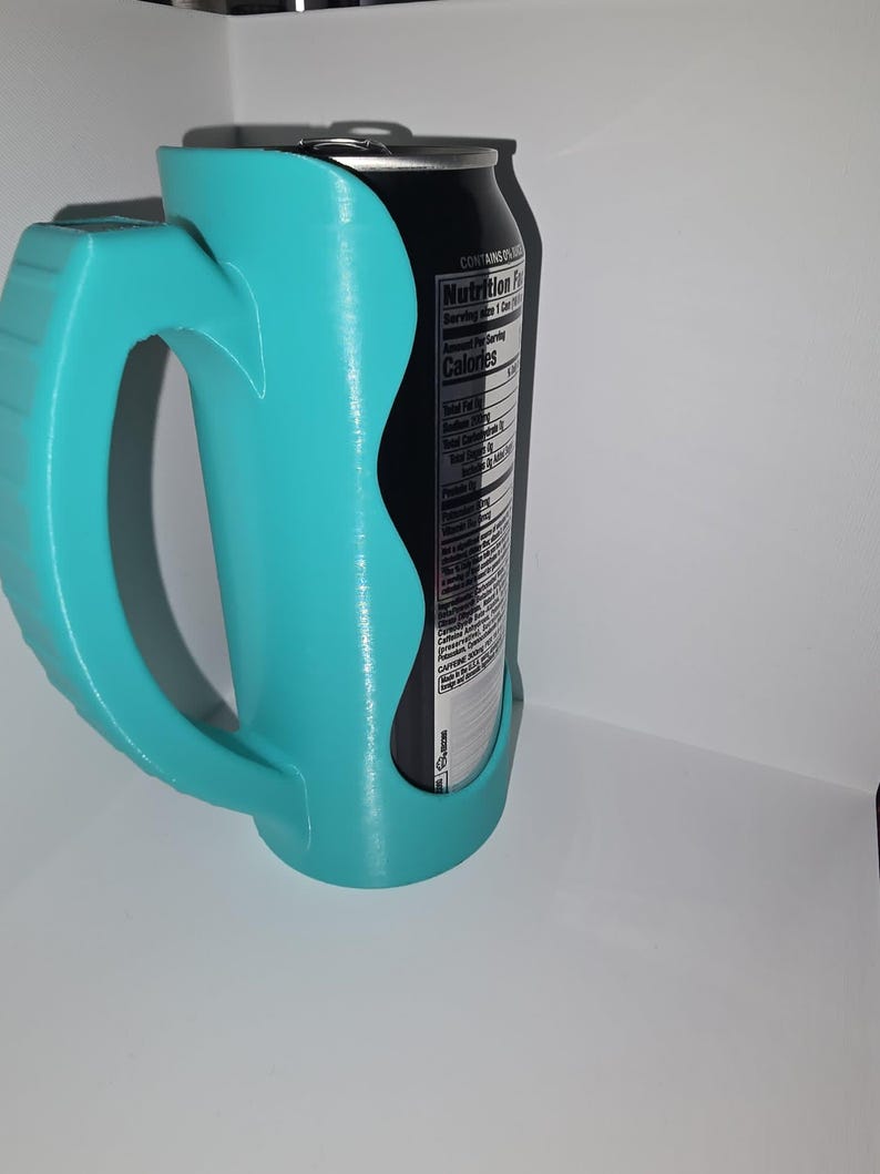 May include: A teal-colored can holder with a handle, designed to hold a standard-sized beverage can. The holder is made of a smooth, possibly 3D-printed material. A black can with a nutrition label is visible inside the holder.
