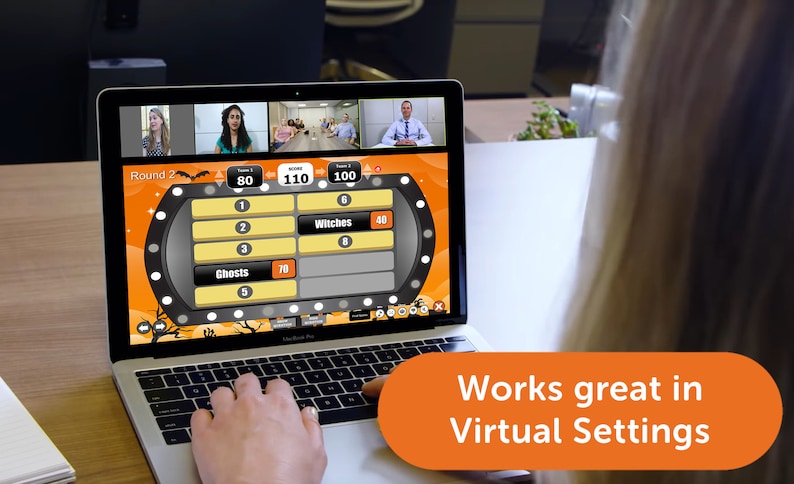 May include: A laptop screen displays a Halloween-themed game show with a scoreboard showing the scores for two teams. The game show is being played virtually with video calls of the players in the top left corner of the screen. The text on the screen reads "Round 2" and "Witches".
