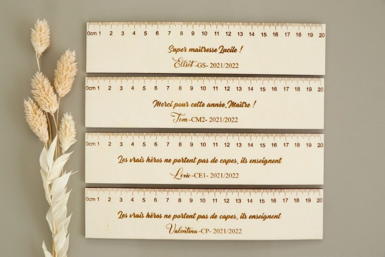 May include: Four wooden rulers with engraved messages and numbers. The top ruler says "Super ma&icirc;tresse Lucille! Elliet-GS-2021/2022". The second ruler says "Merci pour cette ann&eacute;e, Maitre! Tem-CM2-2021/2022". The third ruler says "Les vrais h&eacute;ros ne portent pas de capes, ils enseignent Levic-CE1-2021/2022". The bottom ruler says "Les vrais h&eacute;ros ne portent pas de capes, ils enseignent Valentina-CP-2021/2022".