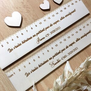 May include: Two wooden rulers with the text "Les vraies h&eacute;ro&iuml;nes ne portent pas de capes, elles enseignent" and the names "Jeana-CP-2020/2021" and "Sandre-CM1-2020/2021" engraved on them. The rulers are decorated with small wooden hearts.