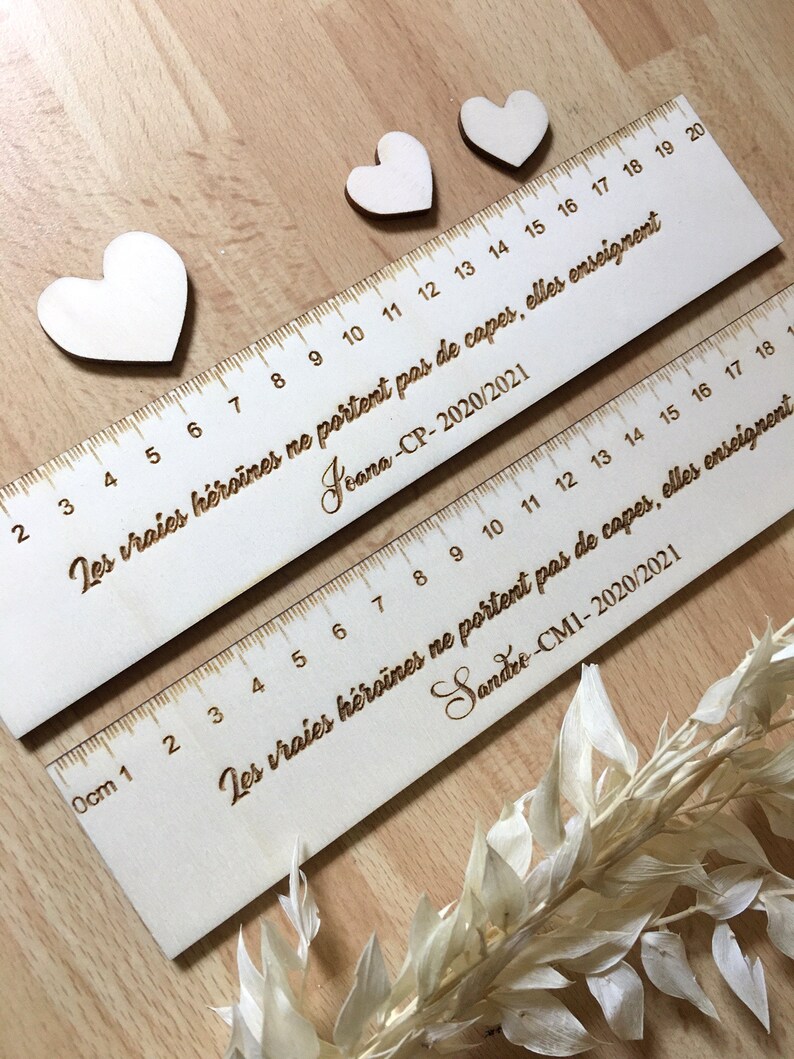 May include: Two wooden rulers with the text "Les vraies h&eacute;ro&iuml;nes ne portent pas de capes, elles enseignent" and the names "Jeana-CP-2020/2021" and "Sandre-CM1-2020/2021" engraved on them. The rulers are decorated with small wooden hearts.