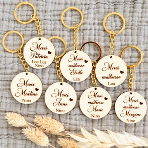 May include: Eight wooden keychains with gold key rings. Each keychain has a round wooden tag with the text "Merci" and a name, such as "Patricia", "Martine", "Anne", "Elodie", "Lou", "Marie", and "Morgane".