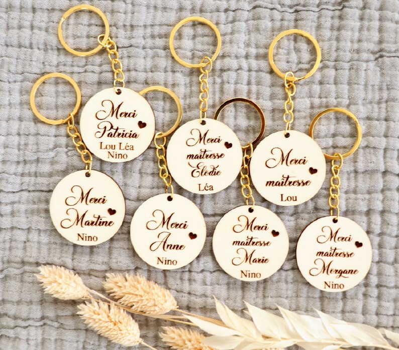 May include: Eight wooden keychains with gold key rings. Each keychain has a round wooden tag with the text "Merci" and a name, such as "Patricia", "Martine", "Anne", "Elodie", "Lou", "Marie", and "Morgane".