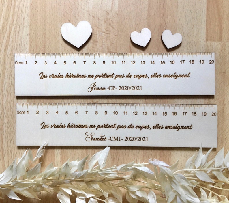 May include: Two wooden rulers with a heart shape at the top. The rulers are marked with centimeters and numbers from 1 to 20. The rulers have the text "Les vraies h&eacute;ro&iuml;nes ne portent pas de capes, elles enseignent" and the names "Jeana-CP-2020/2021" and "Sandee-CM1-2020/2021".