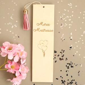 May include: A wooden bookmark with a pink tassel. The bookmark has a gold inscription that reads "Merci Maitresse" with a heart symbol before each word. There are three gold heart-shaped balloons drawn below the inscription.