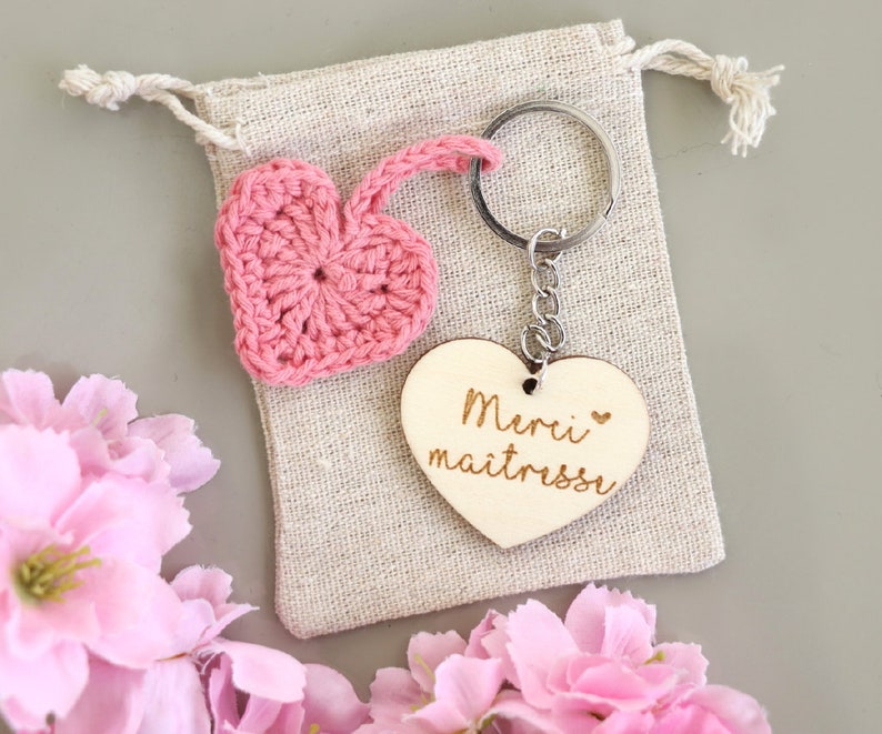 May include: Pink crocheted heart keychain with a wooden heart charm that says "Merci maitresse" in French. The keychain is in a small burlap bag.