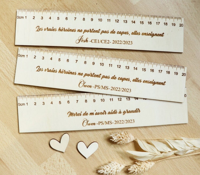 May include: Three wooden rulers with a white background. The rulers are marked with centimeters and numbers from 1 to 20. The rulers have different messages engraved on them: "Les vraies h&eacute;ro&iuml;nes ne portent pas de capes, elles enseignent", "Jesh-CE1/CE2-2022/2023", "Owen-PS/MS-2022/2023", and "Merci de m'avoir aide &agrave; grandir".