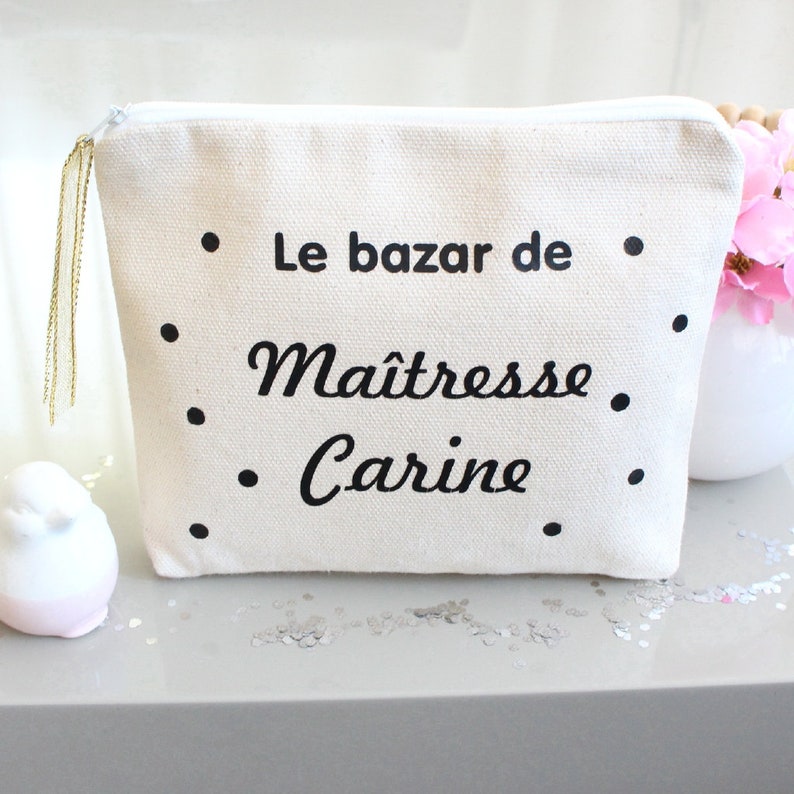 May include: A white canvas makeup bag with a black zipper and a gold tassel. The bag has black polka dots and the text "Le bazar de Maitresse Carine" printed on it.