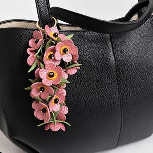 K&ouml;nnte beinhalten: Eine schwarze Ledertasche mit einem floralen Schl&uuml;sselanh&auml;nger. Der Schl&uuml;sselanh&auml;nger zeigt rosa Bl&uuml;ten mit gelben Zentren und schwarzen Akzenten, befestigt an einem goldfarbenen Ring. Die Tasche hat eine strukturierte Oberfl&auml;che und einen oberen Griff.