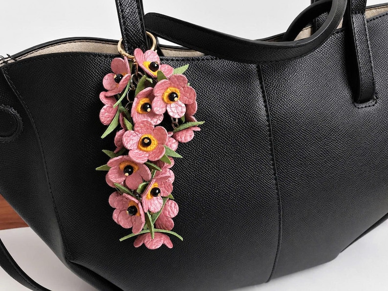 K&ouml;nnte beinhalten: Eine schwarze Ledertasche mit einem floralen Schl&uuml;sselanh&auml;nger. Der Schl&uuml;sselanh&auml;nger zeigt rosa Bl&uuml;ten mit gelben Zentren und schwarzen Akzenten, befestigt an einem goldfarbenen Ring. Die Tasche hat eine strukturierte Oberfl&auml;che und einen oberen Griff.