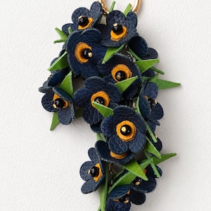 K&ouml;nnte beinhalten: Ein floraler Schl&uuml;sselanh&auml;nger mit marineblauen Lederblumen, gelben Zentren und schwarzen Akzenten, durchsetzt mit gr&uuml;nen Bl&auml;ttern. Der Schl&uuml;sselanh&auml;nger hat einen goldfarbenen Ring zur Befestigung.