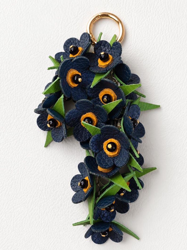 K&ouml;nnte beinhalten: Ein floraler Schl&uuml;sselanh&auml;nger mit marineblauen Lederblumen, gelben Zentren und schwarzen Akzenten, durchsetzt mit gr&uuml;nen Bl&auml;ttern. Der Schl&uuml;sselanh&auml;nger hat einen goldfarbenen Ring zur Befestigung.