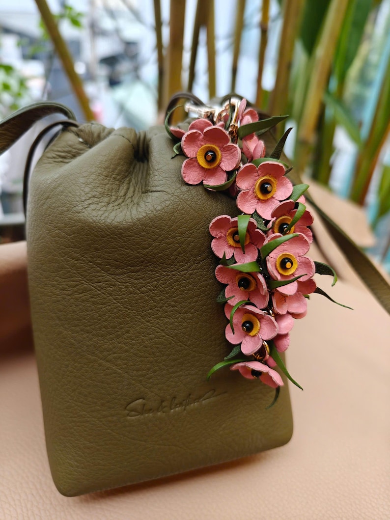 K&ouml;nnte beinhalten: Olivgr&uuml;ner Lederbeutel mit Kordelzug, verziert mit rosa und gelben Blumen. Die Tasche hat einen silberfarbenen Verschluss und einen Lederriemen. Die Tasche ist klein.