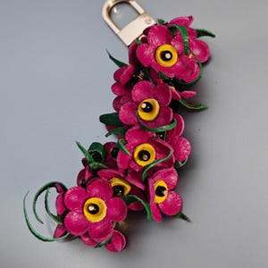 K&ouml;nnte beinhalten: Handgefertigter Leder-Schl&uuml;sselanh&auml;nger mit einer Ansammlung leuchtend magentafarbener Blumen mit gelben Zentren und gr&uuml;nen Akzenten. Die Blumen sind an einem goldfarbenen Metallverschluss befestigt, was ein einzigartiges Accessoire ergibt.