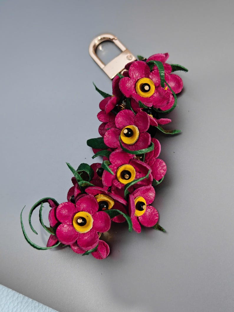 K&ouml;nnte beinhalten: Handgefertigter Leder-Schl&uuml;sselanh&auml;nger mit einer Ansammlung leuchtend magentafarbener Blumen mit gelben Zentren und gr&uuml;nen Akzenten. Die Blumen sind an einem goldfarbenen Metallverschluss befestigt, was ein einzigartiges Accessoire ergibt.