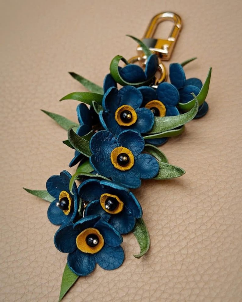 K&ouml;nnte beinhalten: Handgefertigter Leder-Schl&uuml;sselanh&auml;nger mit einem Cluster aus blauen Blumen mit gelben Zentren und schwarzen Akzenten, erg&auml;nzt durch gr&uuml;ne Bl&auml;tter. Der Schl&uuml;sselanh&auml;nger hat einen goldfarbenen Verschluss, ideal f&uuml;r Schl&uuml;ssel oder Taschen.