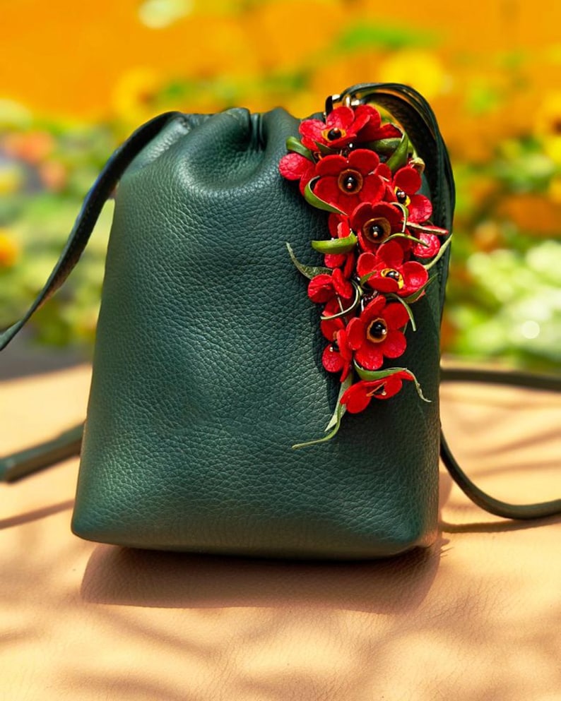 K&ouml;nnte beinhalten: Dunkelgr&uuml;ne Leder-Beuteltasche mit Kordelzug, verziert mit einem Cluster aus roten Blumen und gr&uuml;nen Bl&auml;ttern. Die Tasche hat einen langen Schulterriemen.
