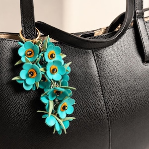 K&ouml;nnte beinhalten: Eine schwarze Ledertasche mit einem t&uuml;rkisfarbenen Blumenanh&auml;nger. Der Anh&auml;nger zeigt mehrere t&uuml;rkisfarbene Blumen mit orangefarbenen Zentren und gr&uuml;nen Bl&auml;ttern, die an einem goldfarbenen Ring befestigt sind. Die Tasche hat ein strukturiertes Design mit einem oberen Griff.