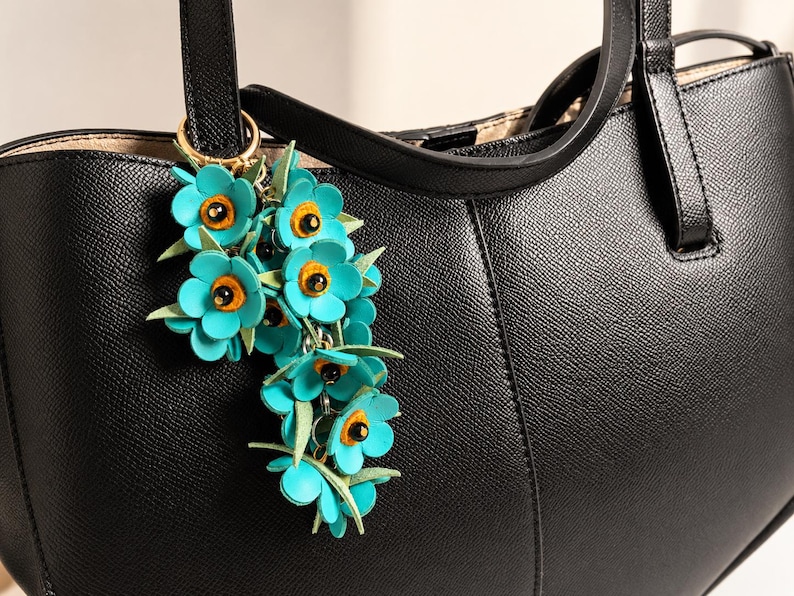 K&ouml;nnte beinhalten: Eine schwarze Ledertasche mit einem t&uuml;rkisfarbenen Blumenanh&auml;nger. Der Anh&auml;nger zeigt mehrere t&uuml;rkisfarbene Blumen mit orangefarbenen Zentren und gr&uuml;nen Bl&auml;ttern, die an einem goldfarbenen Ring befestigt sind. Die Tasche hat ein strukturiertes Design mit einem oberen Griff.