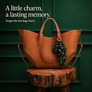 K&ouml;nnte beinhalten: Eine braune Ledertasche mit einem dekorativen Blumenanh&auml;nger. Der Anh&auml;nger zeigt dunkelblaue Blumen mit gelben Zentren und gr&uuml;nen Bl&auml;ttern. Der Text lautet: "Ein kleiner Charme, eine bleibende Erinnerung. Vergissmeinnicht-Taschenanh&auml;nger."