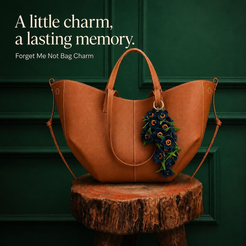 K&ouml;nnte beinhalten: Eine braune Ledertasche mit einem dekorativen Blumenanh&auml;nger. Der Anh&auml;nger zeigt dunkelblaue Blumen mit gelben Zentren und gr&uuml;nen Bl&auml;ttern. Der Text lautet: "Ein kleiner Charme, eine bleibende Erinnerung. Vergissmeinnicht-Taschenanh&auml;nger."
