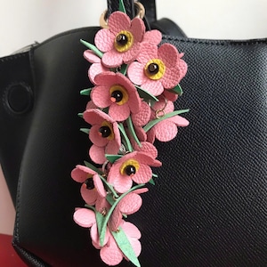 K&ouml;nnte beinhalten: Ein Schl&uuml;sselanh&auml;nger mit rosa Lederblumen, gelben Zentren und gr&uuml;nen Bl&auml;ttern h&auml;ngt an einer schwarzen Handtasche. Die Blumen sind zu einem dekorativen Accessoire zusammengefasst. Der Schl&uuml;sselanh&auml;nger ist an einem goldfarbenen Ring befestigt.
