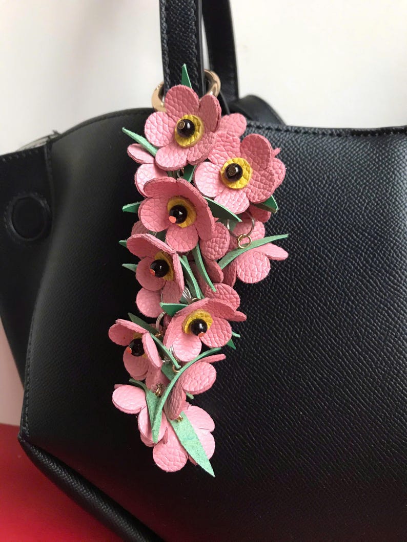 K&ouml;nnte beinhalten: Ein Schl&uuml;sselanh&auml;nger mit rosa Lederblumen, gelben Zentren und gr&uuml;nen Bl&auml;ttern h&auml;ngt an einer schwarzen Handtasche. Die Blumen sind zu einem dekorativen Accessoire zusammengefasst. Der Schl&uuml;sselanh&auml;nger ist an einem goldfarbenen Ring befestigt.