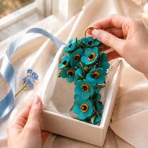 K&ouml;nnte beinhalten: Ein t&uuml;rkisfarbener Blumen-Schl&uuml;sselanh&auml;nger mit schwarzen und gelben Zentren, pr&auml;sentiert in einer wei&szlig;en Geschenkbox. Die Blumen sind in einer Gruppe angeordnet und an einem goldfarbenen Ring befestigt. Ein blaues Band und Vergissmeinnicht sind im Hintergrund.