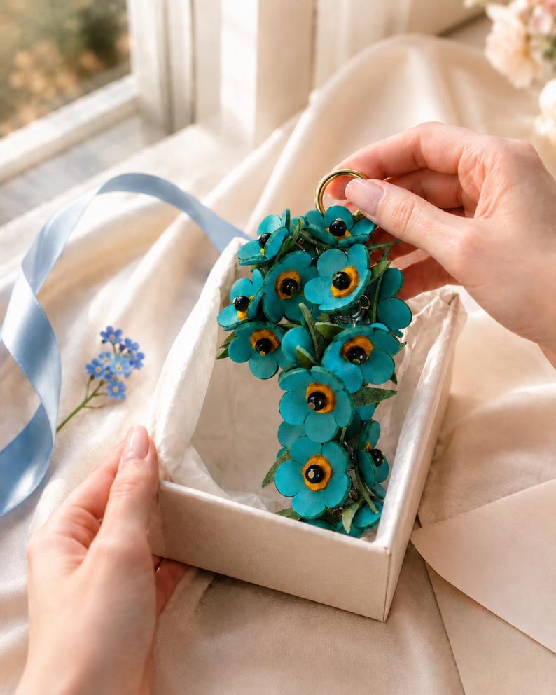 K&ouml;nnte beinhalten: Ein t&uuml;rkisfarbener Blumen-Schl&uuml;sselanh&auml;nger mit schwarzen und gelben Zentren, pr&auml;sentiert in einer wei&szlig;en Geschenkbox. Die Blumen sind in einer Gruppe angeordnet und an einem goldfarbenen Ring befestigt. Ein blaues Band und Vergissmeinnicht sind im Hintergrund.