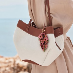 K&ouml;nnte beinhalten: Eine wei&szlig;e und braune Tragetasche mit braunen Lederbes&auml;tzen und -griffen. Die Tasche hat einen floralen Anh&auml;nger. Die Tasche ist aus einem strukturierten Stoff gefertigt. Die Abmessungen der Tasche sind nicht sichtbar.
