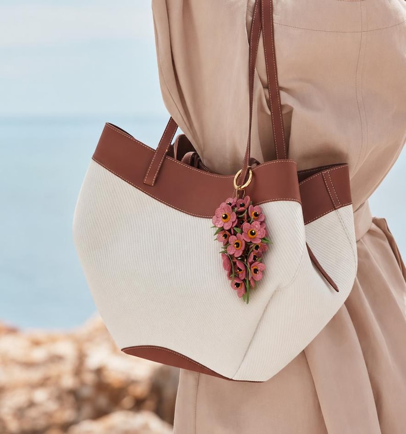 K&ouml;nnte beinhalten: Eine wei&szlig;e und braune Tragetasche mit braunen Lederbes&auml;tzen und -griffen. Die Tasche hat einen floralen Anh&auml;nger. Die Tasche ist aus einem strukturierten Stoff gefertigt. Die Abmessungen der Tasche sind nicht sichtbar.