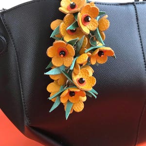 K&ouml;nnte beinhalten: Eine schwarze Handtasche mit einem dekorativen Blumenanh&auml;nger. Der Anh&auml;nger zeigt gelbe Lederblumen mit orangefarbenen Zentren und schwarzen Akzenten, gepaart mit gr&uuml;nen Bl&auml;ttern. Die Tasche ist aus schwarzem Leder.