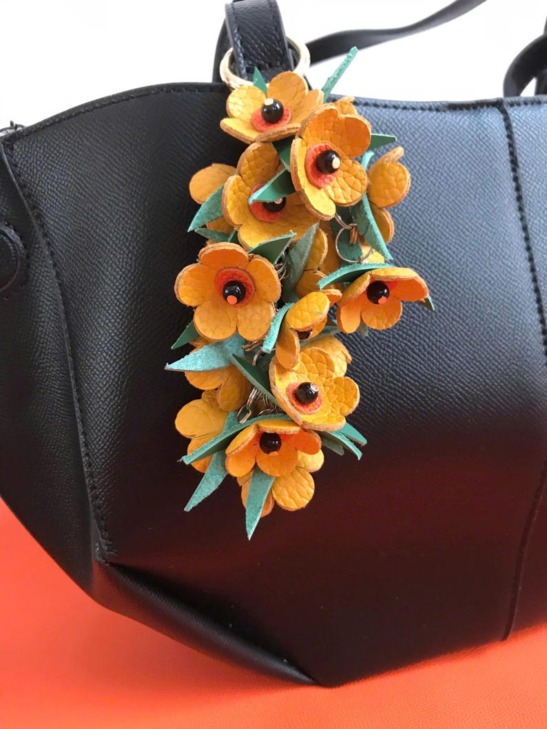 K&ouml;nnte beinhalten: Eine schwarze Handtasche mit einem dekorativen Blumenanh&auml;nger. Der Anh&auml;nger zeigt gelbe Lederblumen mit orangefarbenen Zentren und schwarzen Akzenten, gepaart mit gr&uuml;nen Bl&auml;ttern. Die Tasche ist aus schwarzem Leder.