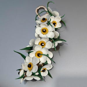 K&ouml;nnte beinhalten: Ein floraler Schl&uuml;sselanh&auml;nger mit wei&szlig;en Lederblumen, gelben Zentren und gr&uuml;nen Bl&auml;ttern. Die Blumen sind an einem goldfarbenen Verschluss befestigt. Das Design ist detailliert, mit einem Fokus auf das nat&uuml;rliche Aussehen der Blumen. Ein dekoratives Accessoire.
