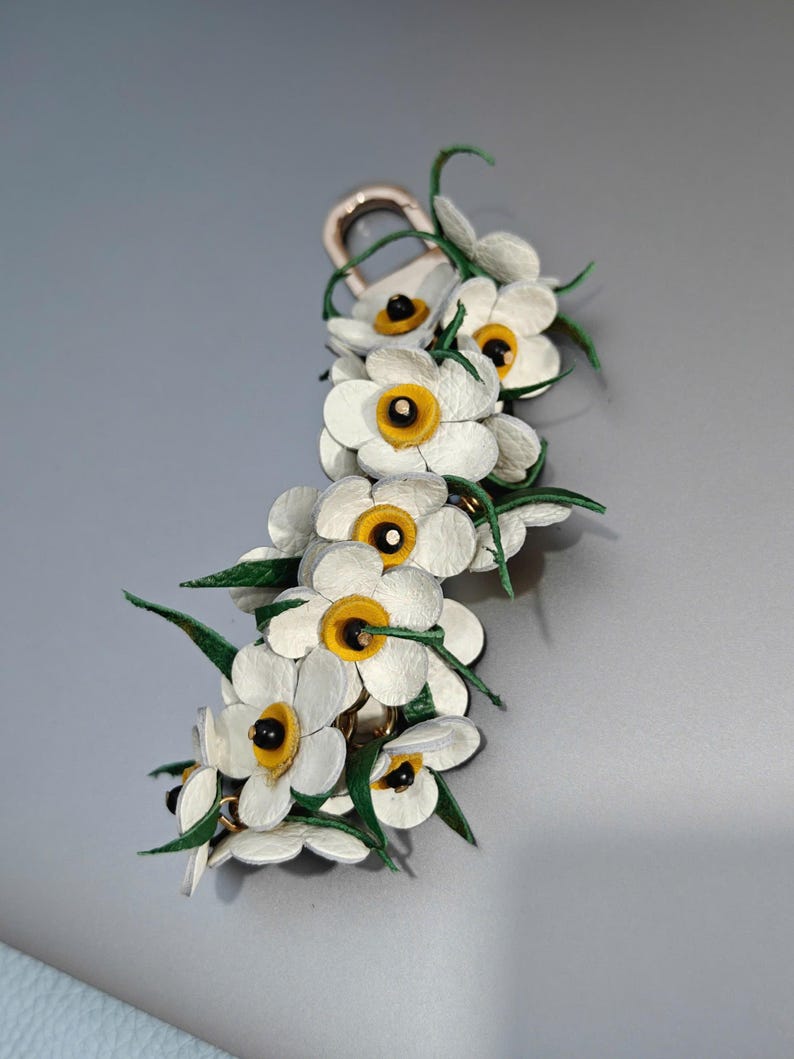 K&ouml;nnte beinhalten: Ein floraler Schl&uuml;sselanh&auml;nger mit wei&szlig;en Lederblumen, gelben Zentren und gr&uuml;nen Bl&auml;ttern. Die Blumen sind an einem goldfarbenen Verschluss befestigt. Das Design ist detailliert, mit einem Fokus auf das nat&uuml;rliche Aussehen der Blumen. Ein dekoratives Accessoire.