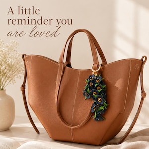 K&ouml;nnte beinhalten: Eine braune Ledertasche mit einem floralen Anh&auml;nger. Die Tasche hat zwei Griffe und einen Schultergurt. Der Anh&auml;nger besteht aus dunkelblauen Blumen mit gelben Zentren. Der Text auf dem Bild lautet: "A little reminder you are loved."