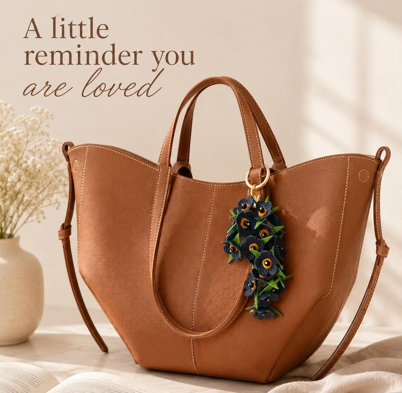 K&ouml;nnte beinhalten: Eine braune Ledertasche mit einem floralen Anh&auml;nger. Die Tasche hat zwei Griffe und einen Schultergurt. Der Anh&auml;nger besteht aus dunkelblauen Blumen mit gelben Zentren. Der Text auf dem Bild lautet: "A little reminder you are loved."