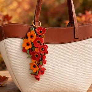 May include: A cream-colored tote bag with a brown leather trim and handles, featuring a decorative hanging charm composed of multiple layered orange, red, and yellow flowers with small black centers attached to a gold-toned metal ring.