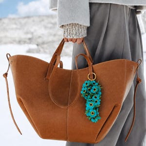 K&ouml;nnte beinhalten: Eine gro&szlig;e, braune Ledertasche mit zwei Henkeln und einem blauen Blumenanh&auml;nger. Die Tasche wird von einer Person getragen, die einen grauen Mantel und eine Hose tr&auml;gt. Der Hintergrund ist eine schneebedeckte Landschaft.