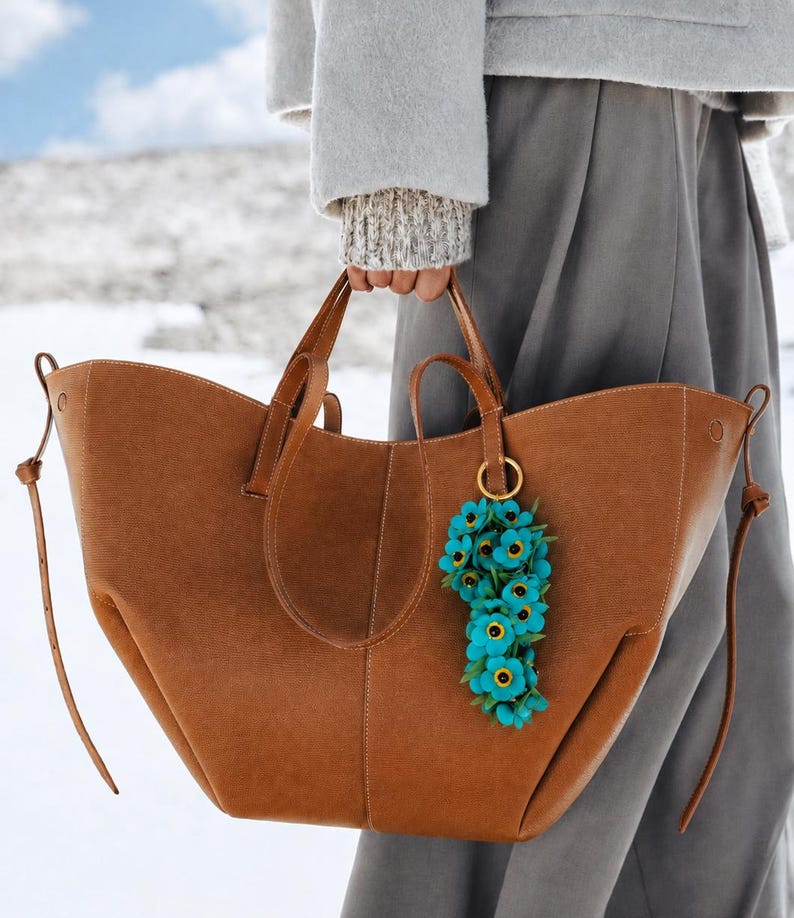 K&ouml;nnte beinhalten: Eine gro&szlig;e, braune Ledertasche mit zwei Henkeln und einem blauen Blumenanh&auml;nger. Die Tasche wird von einer Person getragen, die einen grauen Mantel und eine Hose tr&auml;gt. Der Hintergrund ist eine schneebedeckte Landschaft.