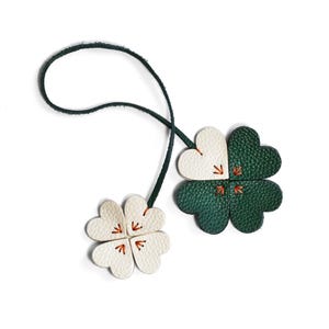 May include: Two leather four-leaf clover charms, one white and one green, connected by a dark green cord. Each clover has orange stitching in the center. The charms are on a white background.