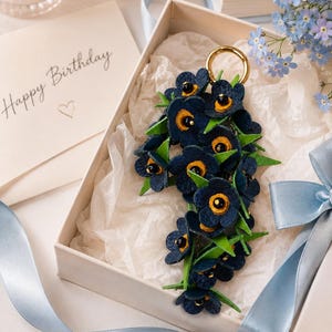 K&ouml;nnte beinhalten: Ein floraler Schl&uuml;sselanh&auml;nger in einer Geschenkbox. Der Schl&uuml;sselanh&auml;nger zeigt dunkelblaue Blumen mit gelben Zentren und gr&uuml;nen Bl&auml;ttern. Oben befindet sich ein goldfarbener Ring. Eine Karte mit "Happy Birthday" befindet sich im Hintergrund, zusammen mit blauem Band und Blumen.