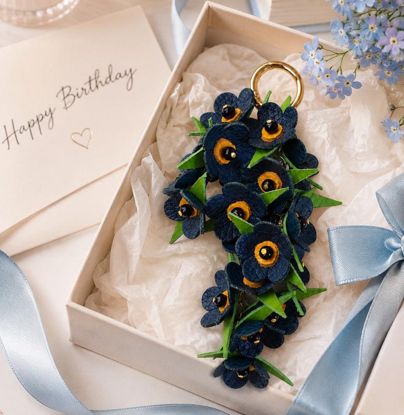 K&ouml;nnte beinhalten: Ein floraler Schl&uuml;sselanh&auml;nger in einer Geschenkbox. Der Schl&uuml;sselanh&auml;nger zeigt dunkelblaue Blumen mit gelben Zentren und gr&uuml;nen Bl&auml;ttern. Oben befindet sich ein goldfarbener Ring. Eine Karte mit "Happy Birthday" befindet sich im Hintergrund, zusammen mit blauem Band und Blumen.