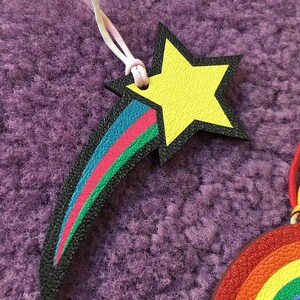 May include: A black leather shooting star with a yellow star at the end. The shooting star has a rainbow stripe design in green, pink, blue, and turquoise. The star is attached to a white and black cord.