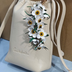 K&ouml;nnte beinhalten: Eine cremefarbene Leder-Umh&auml;ngetasche mit Kordelzugverschluss. Die Tasche ist mit einem Cluster aus wei&szlig;en und gelben Blumen verziert. Sie hat einen langen, d&uuml;nnen Riemen und einen goldfarbenen Verschluss.