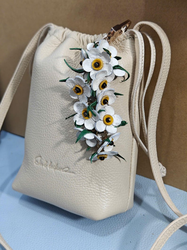 K&ouml;nnte beinhalten: Eine cremefarbene Leder-Umh&auml;ngetasche mit Kordelzugverschluss. Die Tasche ist mit einem Cluster aus wei&szlig;en und gelben Blumen verziert. Sie hat einen langen, d&uuml;nnen Riemen und einen goldfarbenen Verschluss.