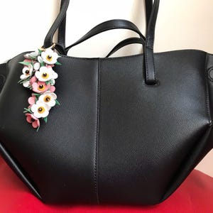 K&ouml;nnte beinhalten: Schwarze Tragetasche mit einem floralen Anh&auml;nger. Die Tasche hat zwei Griffe und einen Schultergurt. Der Anh&auml;nger zeigt wei&szlig;e, rosa und gelbe Blumen. Die Tasche ist aus einem strukturierten Material.